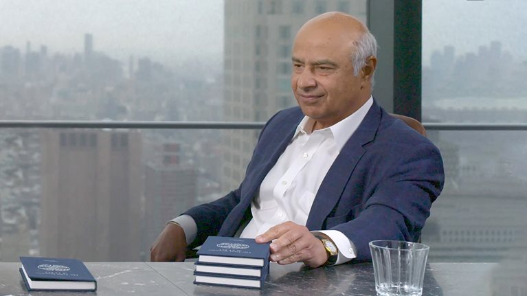 Vik Malhotra seated at a conference room table with his hand on a stack of books. 